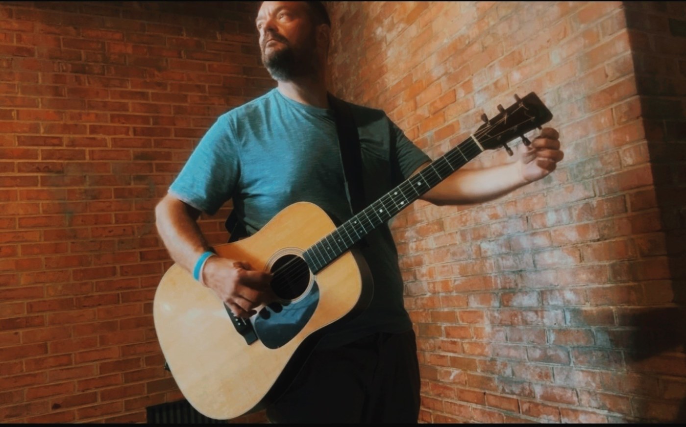 John Leonard playing acoustic guitar against a brick wall